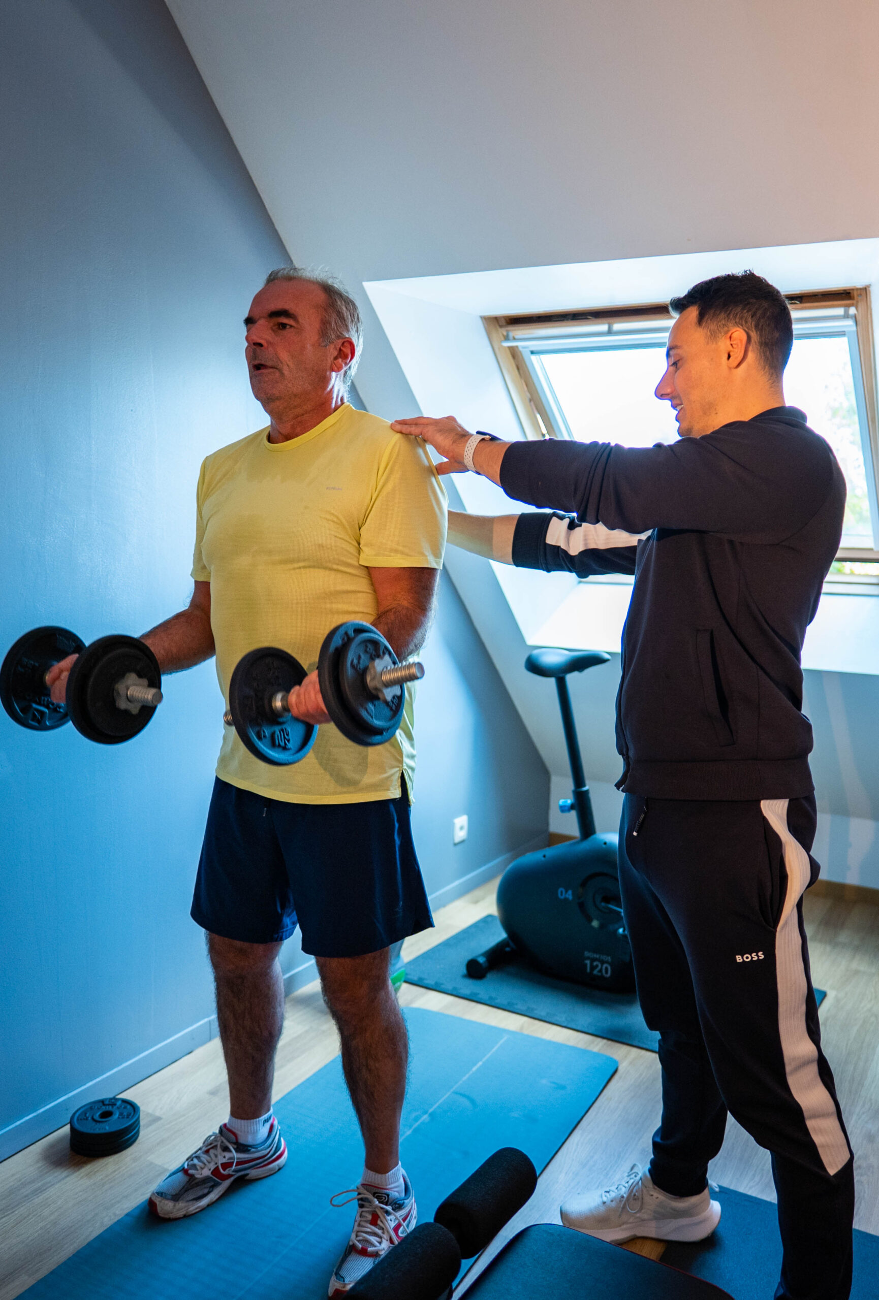 Coach sportif qui entraine un client lors d'une séance de musculation avec haltères, dans une salle d'entraînement privée. Dans le home gym du client, le coach apporte des instructions pour améliorer la sécurité de l'exécution des mouvements.