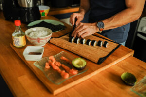 Le coach nutritionnel et sportif Alexis découpe des makis fait maison. On voit au premier rang du riz à sushis, du vinaigre pour riz, du saumon, de l'avocat, de la sauce soja. Il découpe ses makis sur son plan de travail, à côté de feuilles d'algues.